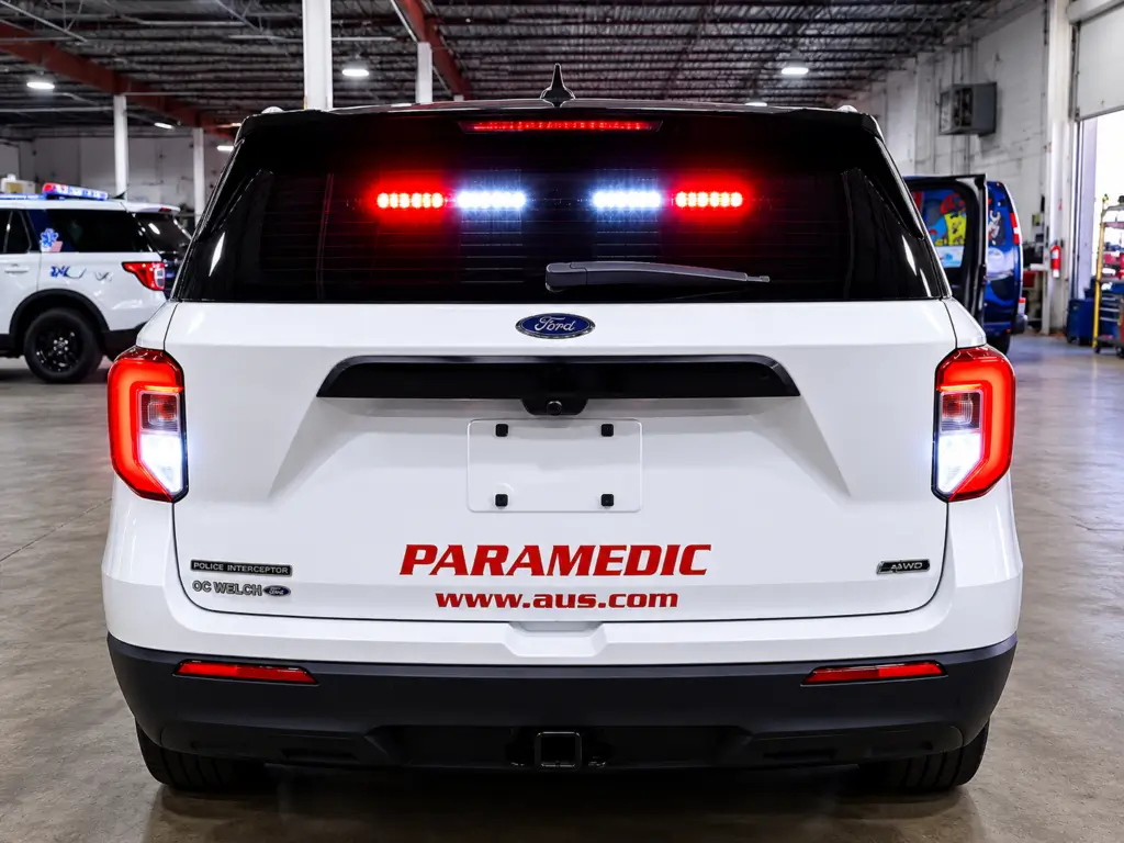 Rear view of a white Ford Police Interceptor Utility vehicle equipped with flashing red and white emergency lights, custom paramedic markings, and public safety upfitting features inside a professional emergency vehicle upfitting facility.