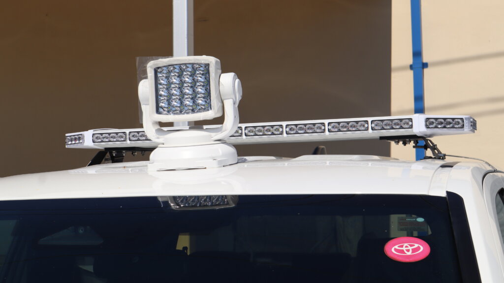 Close-up of a white utility vehicle equipped with a roof-mounted LED warning light bar and remote-controlled spotlight, designed for fleet, public works, emergency response, and roadside safety operations.