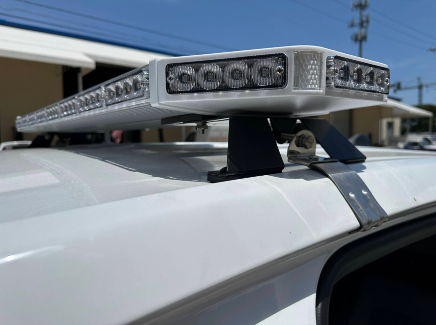 Close-up of a low-profile LED emergency light bar mounted on the roof of a white fleet vehicle, featuring clear warning light modules and durable mounting brackets for public safety, utility, and service vehicle applications.