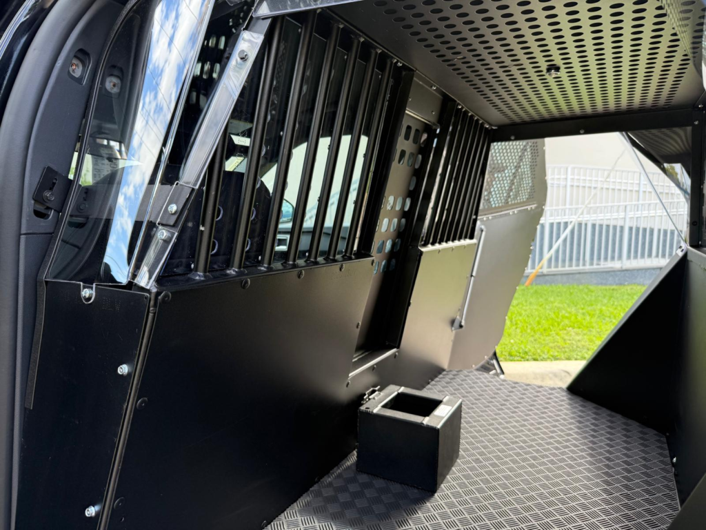 Interior view of a police vehicle prisoner transport compartment featuring reinforced steel partitions, barred security panels, durable diamond-pattern flooring, and a secure rear holding area designed for safe detainee transport.