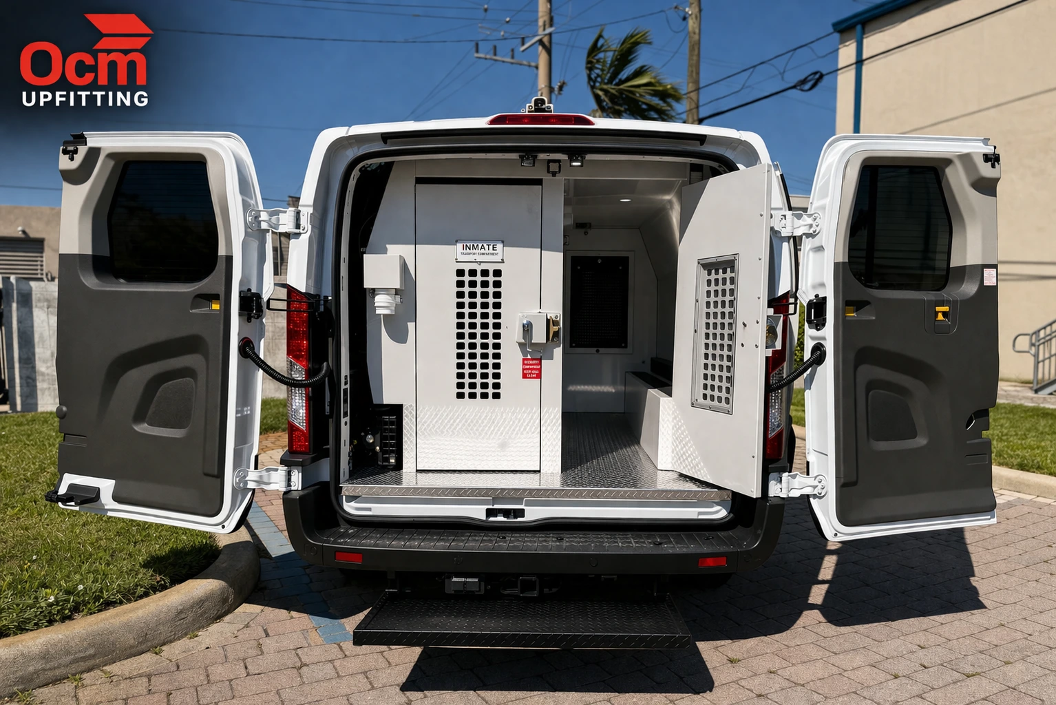 A wide-angle photograph from the rear of a white Ford Transit prisoner transport van with all its rear doors wide open. The interior features a heavy-duty, secure metal containment cage dividing the cargo area into smaller individual cells, complete with mesh windows and heavy latches, against a backdrop of an industrial park with palm trees under a bright blue sky.